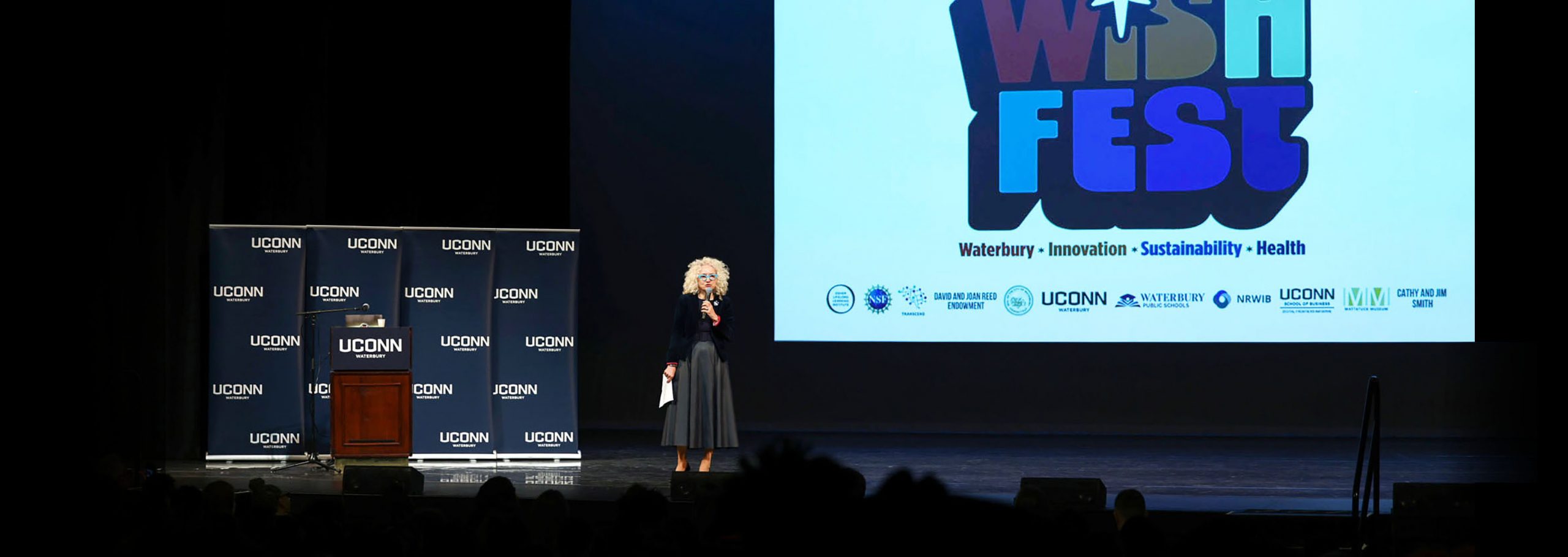 UConn president Radenka Maric speaks on stage with a slide showing the WISHfest logo and sponsors